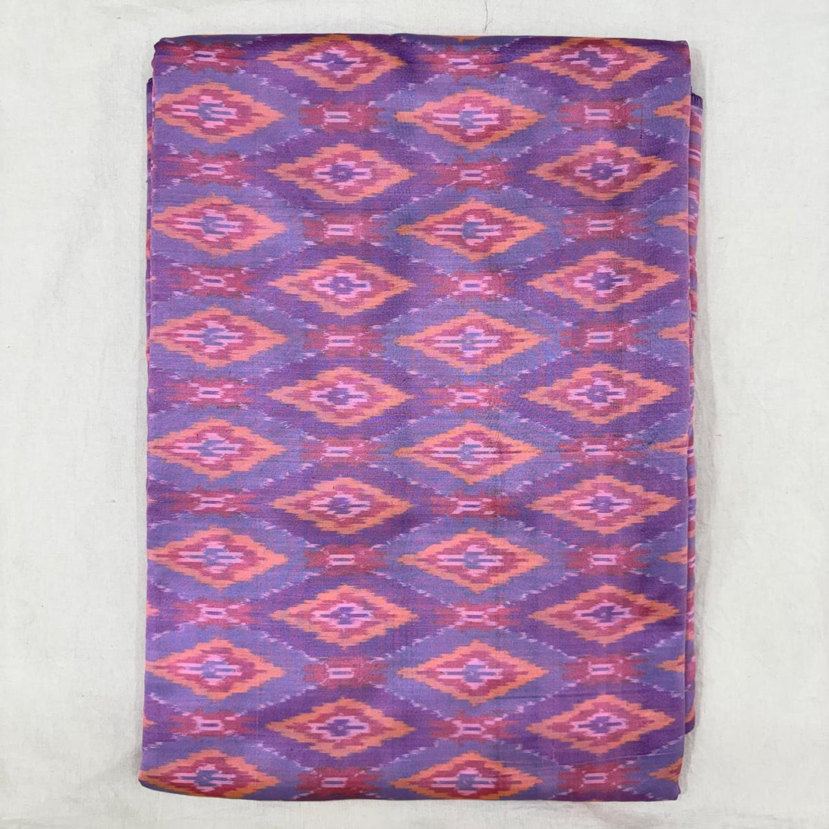 Folded lavender violet pure silk Ikat fabric featuring geometric diamond lozenge motifs in coral peach and lilac with Silk Mark certification.