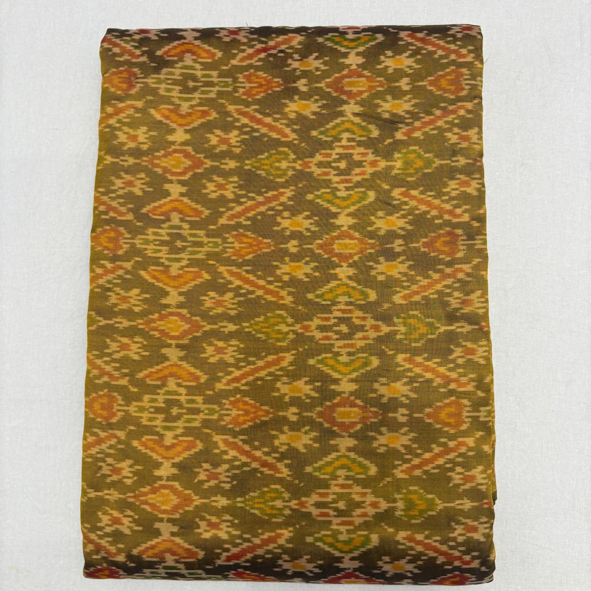Folded earthy olive green pure silk Ikat fabric featuring traditional geometric diamond and amber rust motifs with Silk Mark certification.