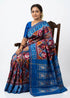A beautiful traditional Indian woman sitting on a carved wooden chair against a stark white background. She is elegantly draped in a vibrant royal blue and maroon Patola-inspired Ikat silk saree featuring intricate geometric patterns. The saree has a prominent royal blue pallu detailed with traditional woven elephant motifs, and she is wearing a matching solid royal blue silk blouse.