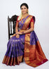 Indian woman wearing a royal blue semi Dharmavaram silk saree with red zari border, sitting gracefully on an antique wooden chair against a plain white background.