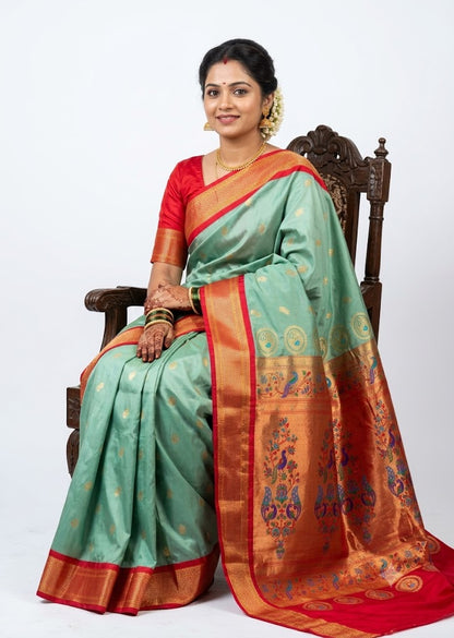 A smiling Indian woman seated gracefully on a carved wooden chair, wearing an elegant sea green Semi Gadwal Paithani saree with a rich gold and red zari border and a matching red blouse, set against a solid white background.