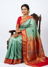 A smiling Indian woman seated gracefully on a carved wooden chair, wearing an elegant sea green Semi Gadwal Paithani saree with a rich gold and red zari border and a matching red blouse, set against a solid white background.