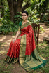 A graceful Indian woman seated on a wooden chair in a lush outdoor garden, wearing a crimson red semi-Dharmavaram silk saree with gold zari checks, a rich deep green contrast border, and an ornate pallu, styled with antique gold jewelry.