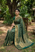 A graceful Indian woman seated on a wooden chair in a lush garden, wearing a rich emerald green semi-Dharmavaram silk saree with intricate gold zari checks and a heavy traditional border, styled with antique gold jewelry.