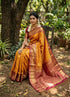 Traditional Indian woman seated gracefully wearing a vibrant yellow-gold and maroon-red Semi Dharmavaram Silk Saree with intricate gold zari brocade borders, elaborate geometric pallu patterns, and tassels in a lush heritage garden.