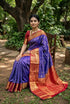 A graceful Indian woman seated on a wooden chair in a garden, wearing a royal blue and deep red semi-Dharmavaram silk saree with intricate gold zari checks, a heavy red border, and an ornate pallu, paired with traditional antique-gold jewelry.