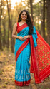 Smiling Indian woman outdoors draped in a sky blue ikat silk saree paired with an intricate red and yellow patterned pallu