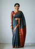 Woman wearing a traditional deep teal blue Kuppadam Pattu saree with wavy silver and copper zari stripes, copper zari border, and contrast maroon red floral brocade pallu and blouse.