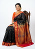 A graceful Indian woman seated on a carved wooden chair wearing a traditional black silk saree with a red and gold zari border, an ornate peacock pallu, and an orange blouse, against a white studio backdrop.