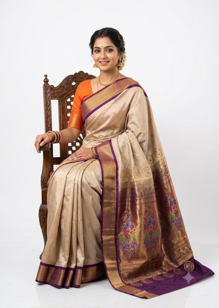 A beautiful traditional Indian woman seated on a carved wooden chair wearing a champagne gold silk saree with a deep purple and gold zari border, an ornate peacock pallu, and an orange blouse, against a white studio backdrop.