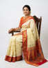 A beautiful traditional Indian woman seated on a carved wooden chair wearing a cream silk saree with a red and gold zari border, an ornate peacock pallu, and an orange blouse, against a white studio backdrop.