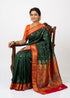 Smiling traditional Indian woman sitting on a wooden chair wearing a dark green silk saree with a red and gold zari border and peacock motifs, against a plain white background.