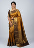 A beautiful traditional Indian woman smiling and posing in a golden yellow and dark brown Dharmavaram silk saree with intricate gold zari work, featuring a matching brown blouse, gold jewelry, and jasmine flowers in her hair against a light gray backdrop.