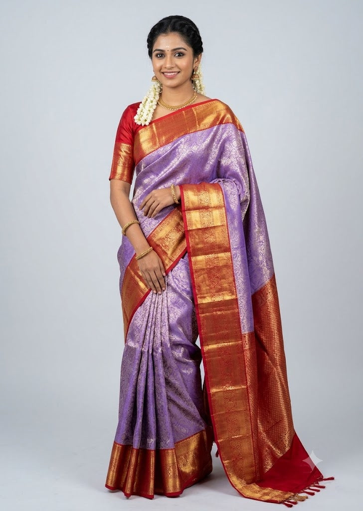 A beautiful traditional Indian woman smiling and posing in a lavender and red Dharmavaram silk saree with intricate gold and silver zari work, featuring a matching red blouse and gold jewelry against a light gray backdrop.