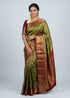 A beautiful traditional Indian woman smiling and posing in an olive green and maroon Dharmavaram silk saree with elaborate gold zari work, featuring a matching maroon blouse, traditional gold jewelry, and jasmine flowers in her hair against a light gray backdrop.