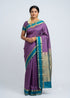 Full-length view of an Indian woman in a purple Kanchi silk saree with teal blue border, gold jewelry, and jasmine flowers, against a light grey background.