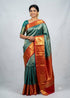An Indian woman in a traditional pose wearing a beautiful teal-green and orange semi-Dharmavaram silk saree with intricate gold zari work, standing against a light grey studio background.