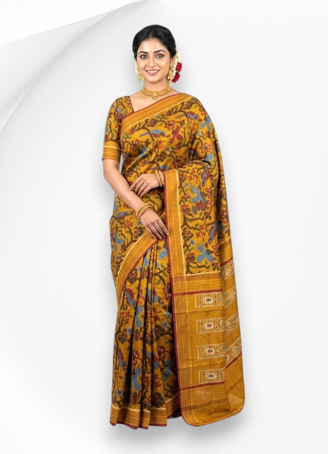 A smiling woman wearing a traditional handwoven mustard yellow Ikkat Patola silk saree featuring intricate floral and bird motifs, accessorized with gold jewelry against a white background.